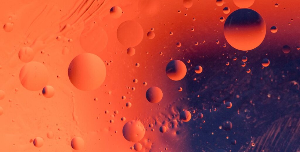 Macro shot of backlit water-oil emulsion over colored background. Tiny gas bubbles inside larger oil bubbles inside water base. Drops of water and scratches on outer surface of the plastic container.
