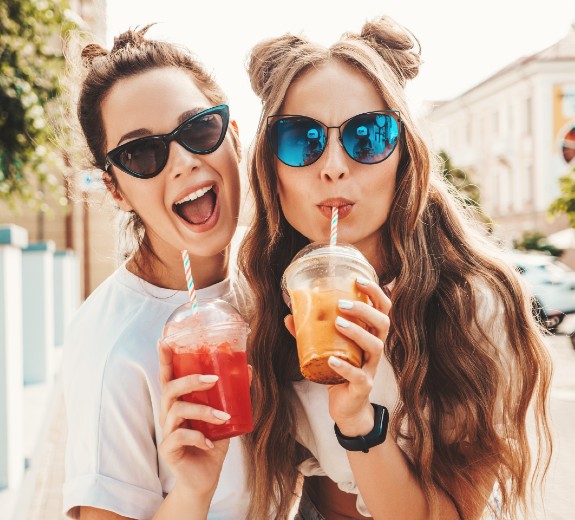 Two young beautiful smiling hipster females in trendy summer clothes. Carefree women posing outdoors. Positive models holding and drinking fresh cocktail drinks in plastic cups with straws.